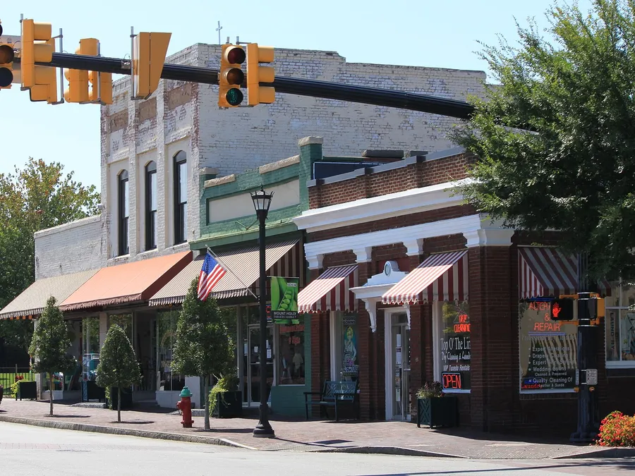 Brick storefronts with striped awnings in downtown Mount Holly, NC, with a farmers market banner on a lamppost and traffic lights overhead